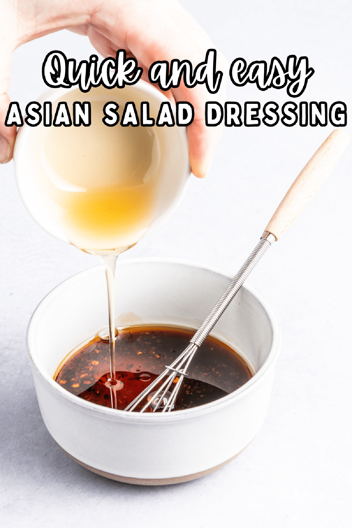 A white bowl of dark brown colored Asian salad dressing with a small whisk inside the bowl; a hand is pouring rice wine vinegar from a second bowl into the dressing.