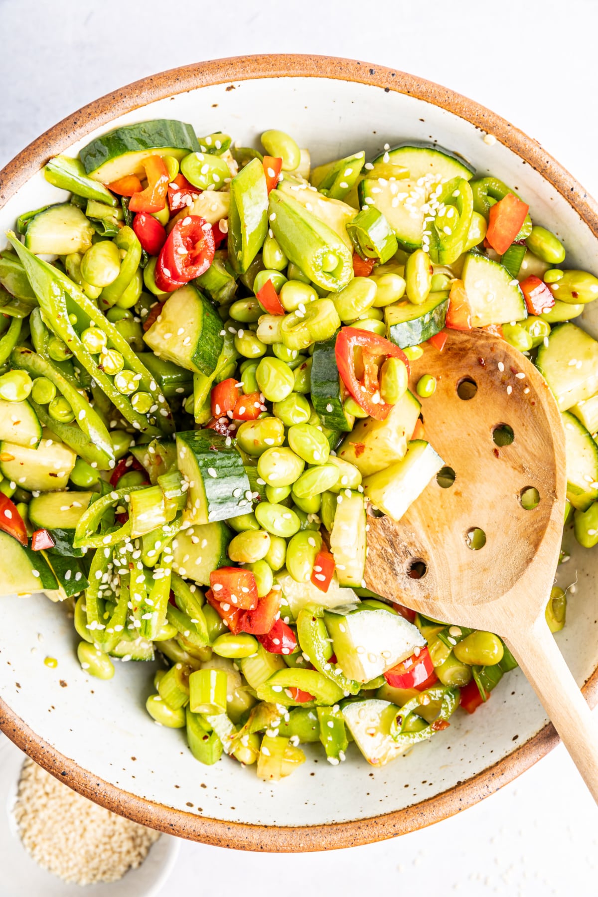 Overhead view of edamame cucumber salad in a bowl with a wooden slotted spoon: salad has edamame, cucumber, snap peas, red bell pepper, green onions, chili peppers and sesame seeds.