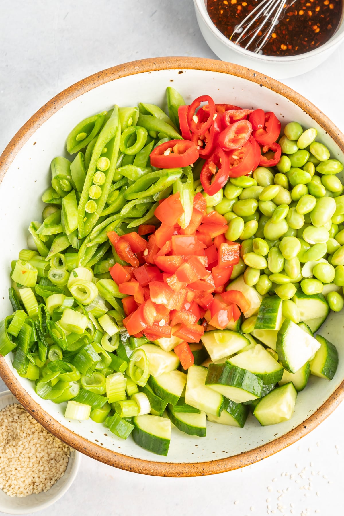 Overhead view of edamame cucumber salad in a bowl before tossing together: salad has edamame, cucumber, snap peas, red bell pepper, green onions, chili peppers and sesame seeds.