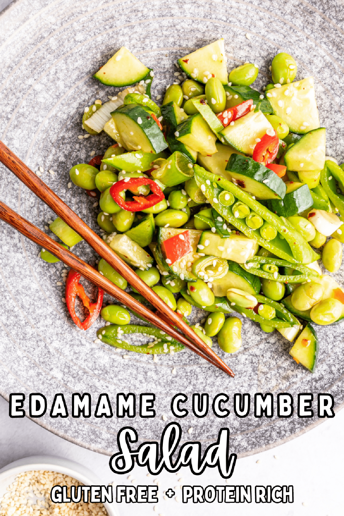 Overhead view of a single serving of Asian cucumber salad in a grey bowl with wooden chopsticks: salad has edamame, cucumber, snap peas, red bell pepper, green onions, chili peppers and sesame seeds.