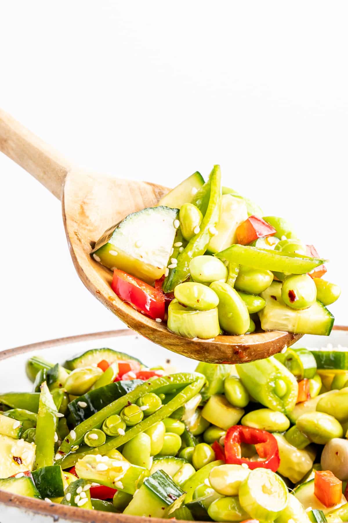 A wooden spoon full of edamame cucumber salad over a bowl of the salad: salad has edamame, cucumber, snap peas, red bell pepper, green onions, chili peppers and sesame seeds.