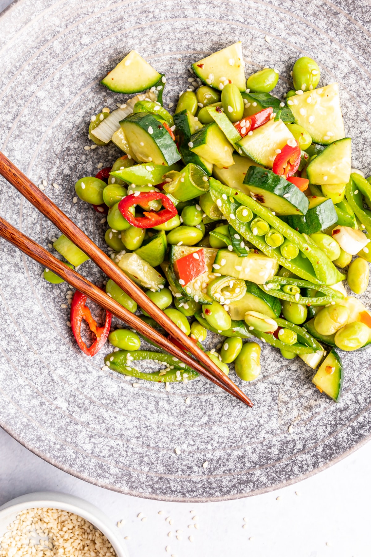Overhead view of a single serving of edamame cucumber salad in a grey bowl with wooden chopsticks: salad has edamame, cucumber, snap peas, red bell pepper, green onions, chili peppers and sesame seeds.