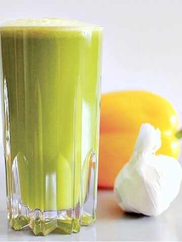 Side view of bright green garlic apple juice in a tall cut crystal glass. juice has light green foam on top, and there is a yellow bell pepper and a white bulb of garlic sitting beside the glass of juice.