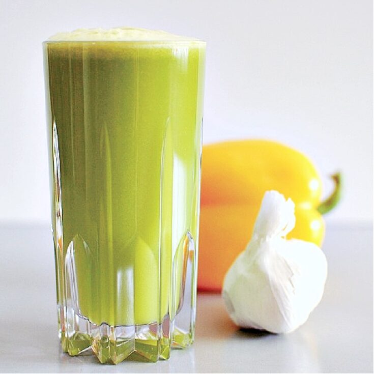 Side view of bright green garlic apple juice in a tall cut crystal glass. juice has light green foam on top, and there is a yellow bell pepper and a white bulb of garlic sitting beside the glass of juice.
