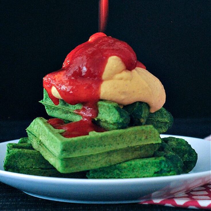 Bright green spinach waffles stacked three high on a white plate, topped with orange colored sriracha whip cream and bright red sriracha sauce.