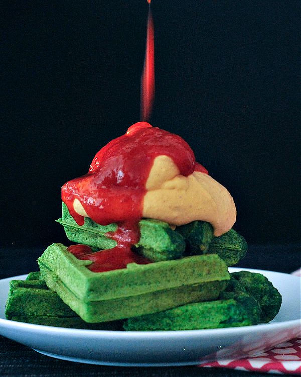 Bright green spinach waffles stacked three high on a white plate, topped with orange colored sriracha whip cream and bright red sriracha sauce.