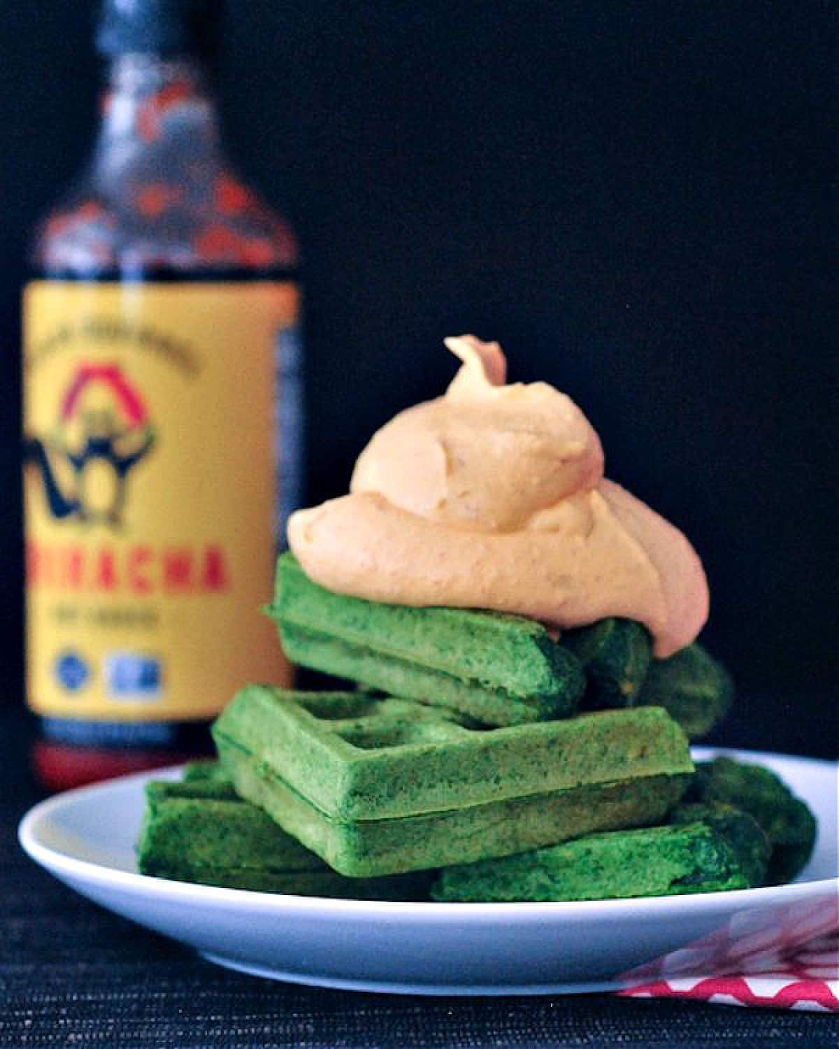 Bright green spinach waffles stacked three high on a white plate, topped with orange colored sriracha whip cream. Sriracha hot sauce bottle in background.