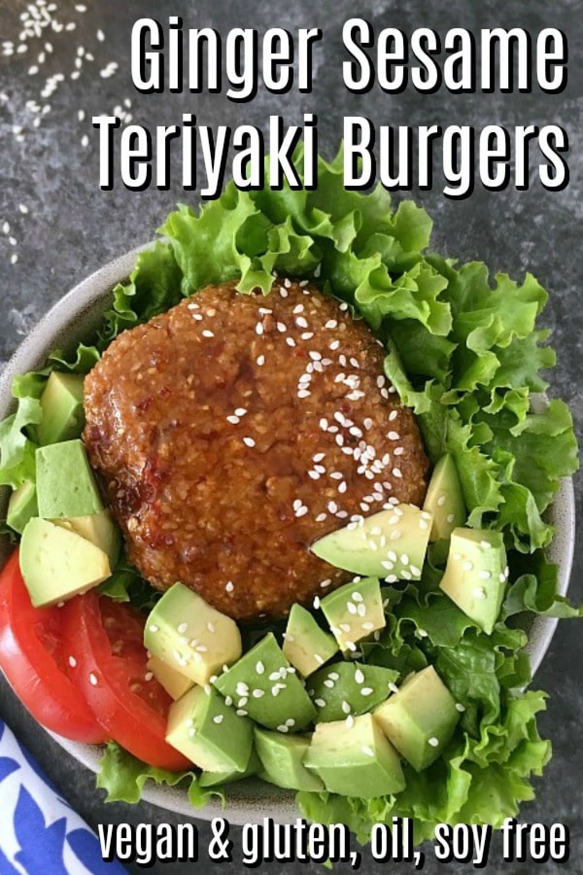 Overhead view of a ginger sesame teriyaki burger sitting on a bed of curly lettuce, tomato slices, and cubed avocado. The whole dish is topped with white sesame seeds, and a fork on a blue and white napkin sits alongside.