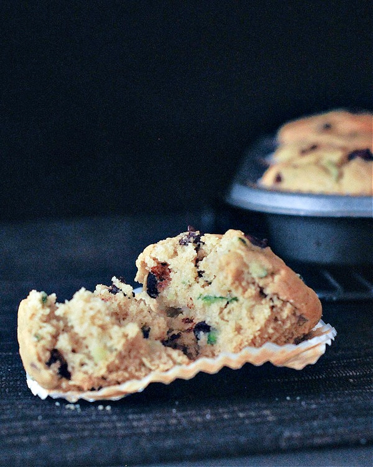 One chocolate chip zucchini muffin cut in half, sitting on a dark placemat against a black background, with a muffin tin full of muffins in background.