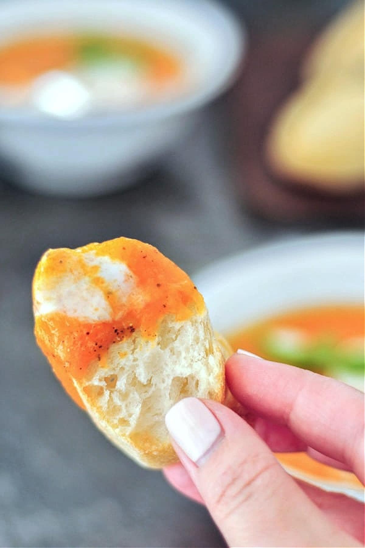 A hand holding a slice of crusty bread dipped into bright orange sweet potato soup.