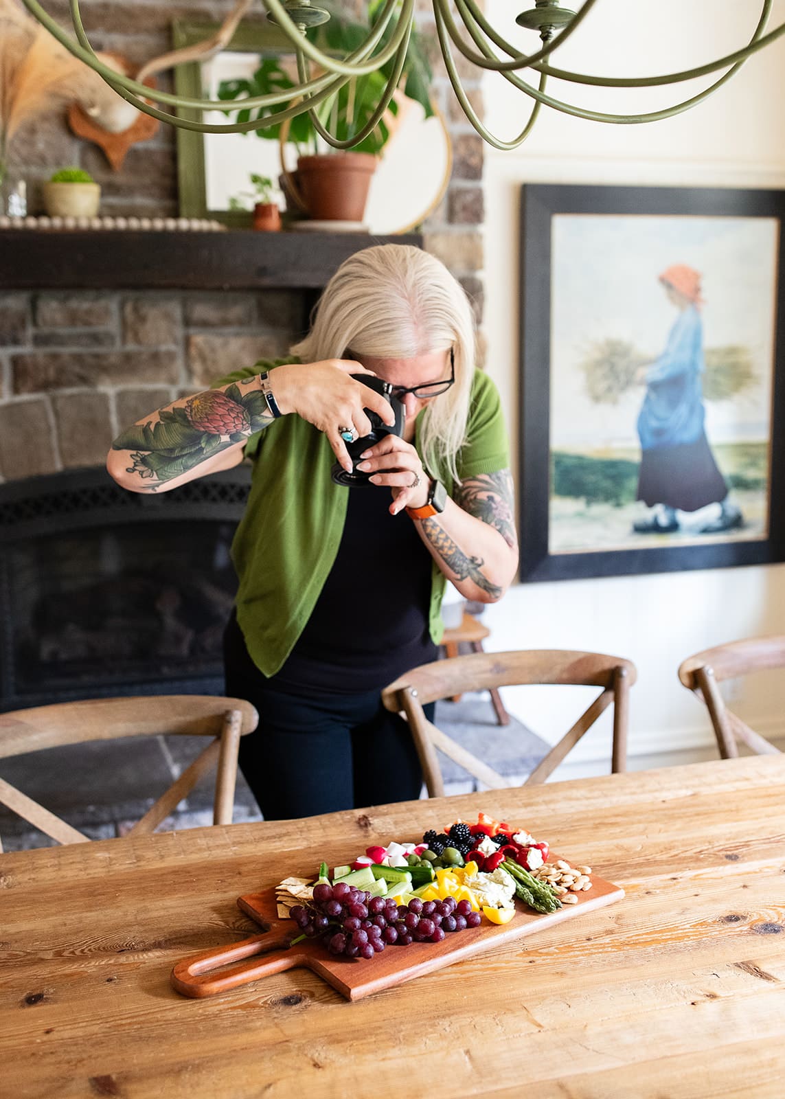 Kristina is standing over a table, pointing her camera at a colorful snack board of fruit, veggies, crackers, nuts, and vegan cheeses.
