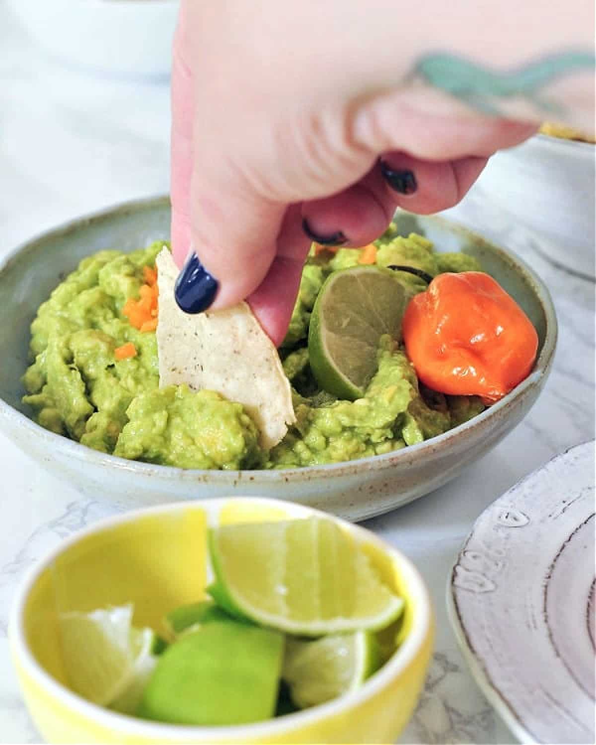 A hand dipping a chip into a bowl of mango habanero guacamole. A smaller bowl of lime wedges on the side.