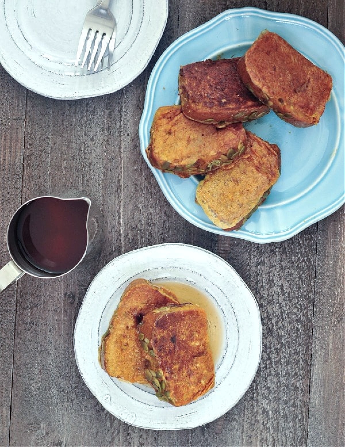 Overhead view of pumpkin bread French toast - a plate full of slices, one plate with two slices and syrup, extra plates and forks, and a silver pitcher of warm syrup.