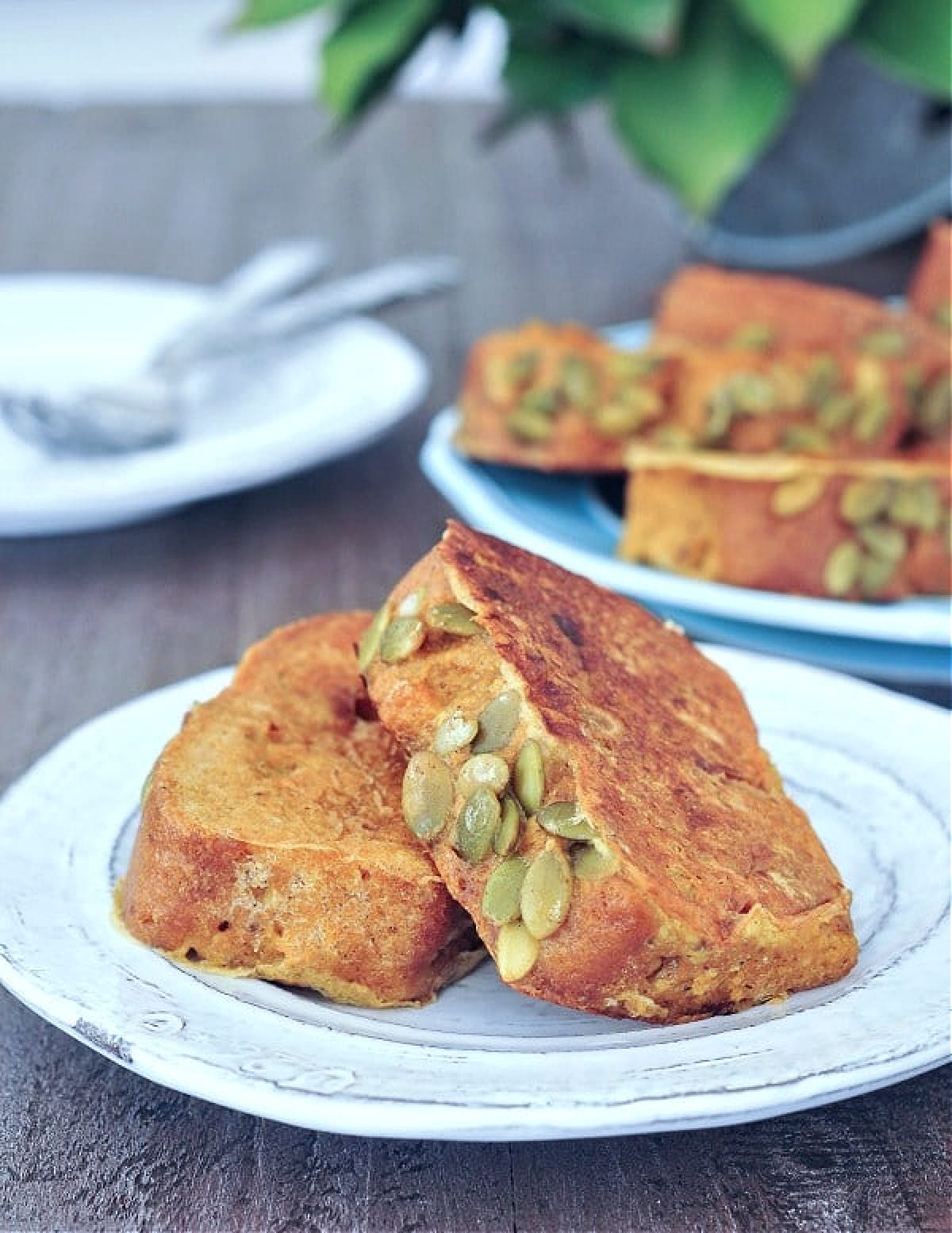 Two thick slices of pumpkin bread French Toast on a plate, a platter full of French toast and a stack of plates and forks is blurred in background.