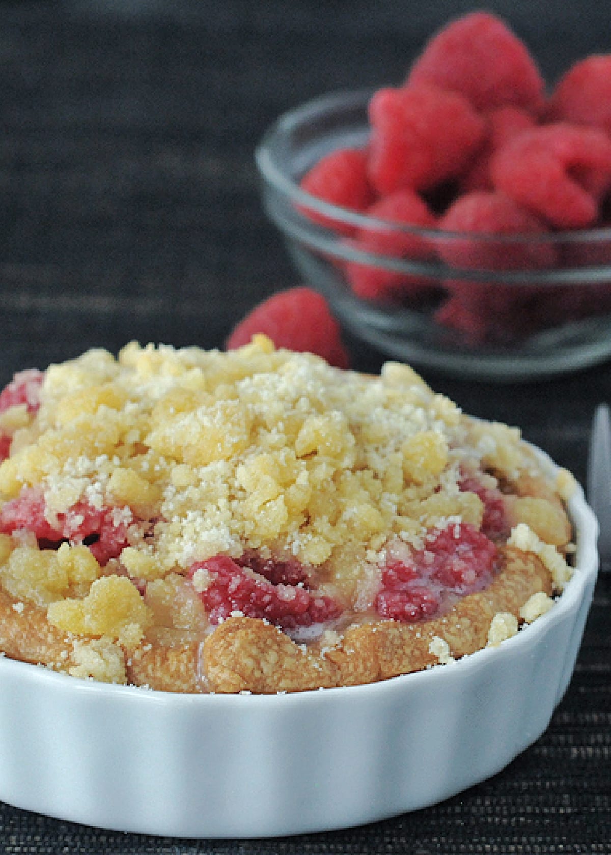 Raspberry crumble pie in a small white ramekin, with a small glass bowl of fresh raspberries in background.