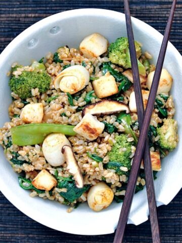 Overhead view of sesame scallop fried rice in a bowl with chopsticks: vegan "scallops" made from hearts of palm, sliced shiitake mushroom, broccoli trees, sliced green onion, golden fried rice.