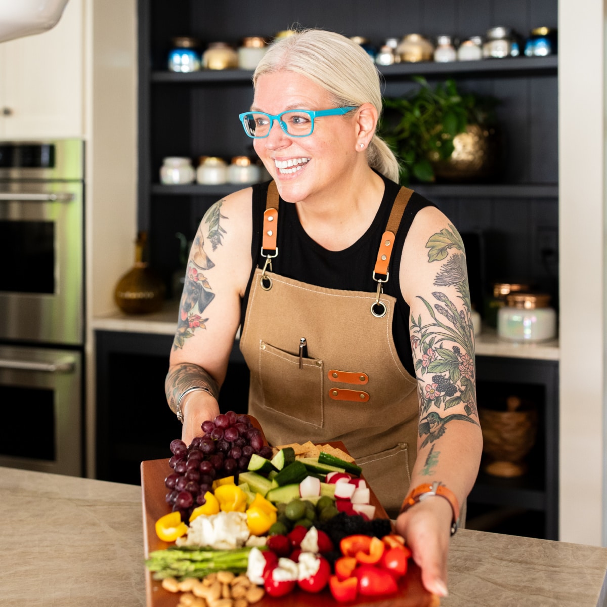Kristina, founder of spabettie.com, is standing at a stone countertop holding a large wooden board of veggies, fruits, cheeses and crackers. She is wearing a tan chefs apron over a black sleeveless top, showing tattooed arms.