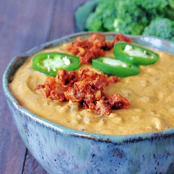 Spicy Vegan Chorizo Queso in a bowl, garnished with fresh sliced jalapeno. Broccoli trees for dipping in background.