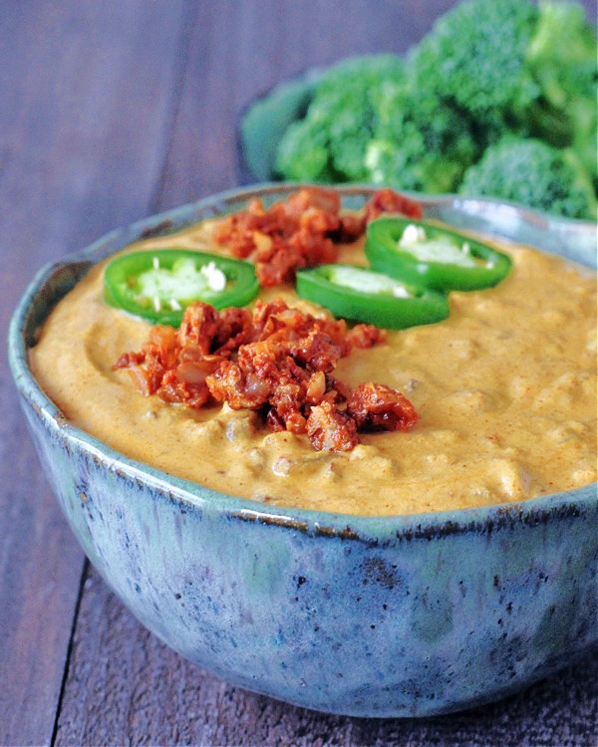 Spicy Vegan Queso in a bowl, garnished with fresh sliced jalapeno. Broccoli trees for dipping in background.