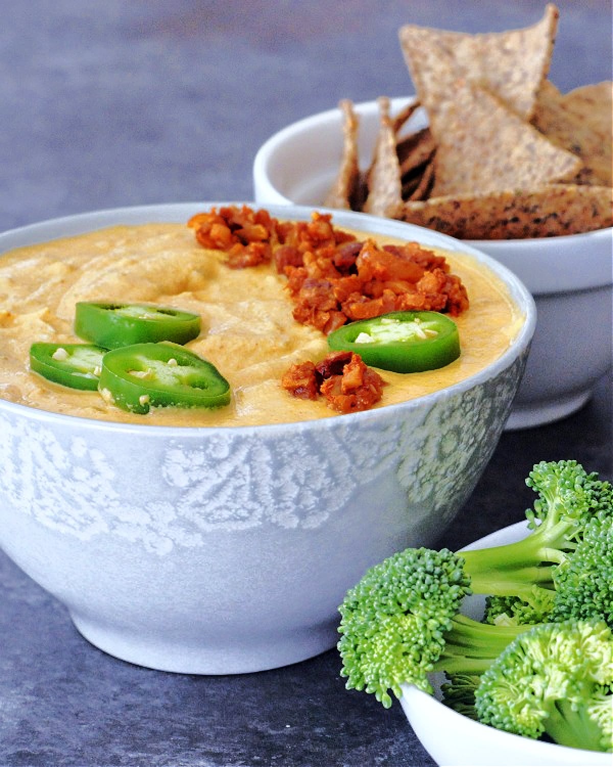 Spicy vegan chorizo queso in a grey bowl, garnished with sliced fresh jalapeno and chorizo crumbles, with another bowl of chips in the background, and broccoli trees for dipping in front of the bowl of queso.