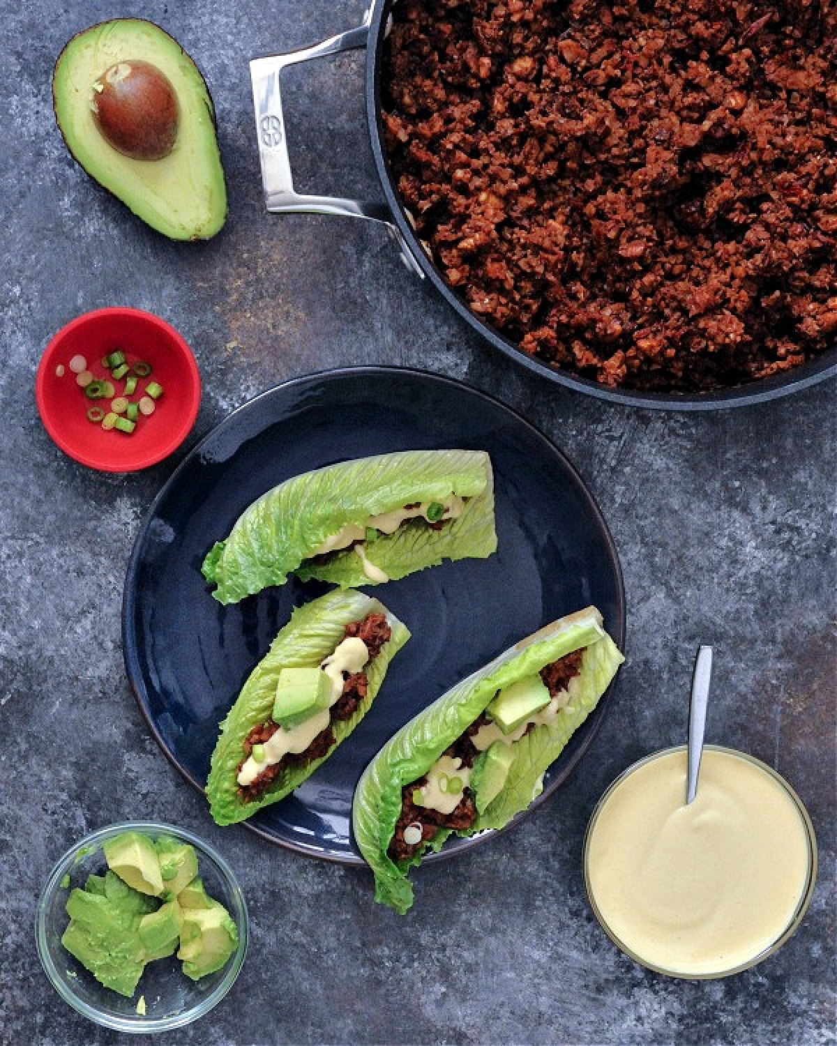 Overhead view of vegan chorizo sausage in a skillet, next to lettuce wraps with the chorizo, avocado slices and dairy free cheese sauce. A small bowl of cheese sauce, a half avocado, and a smaller bowl of sliced avocado on the side.