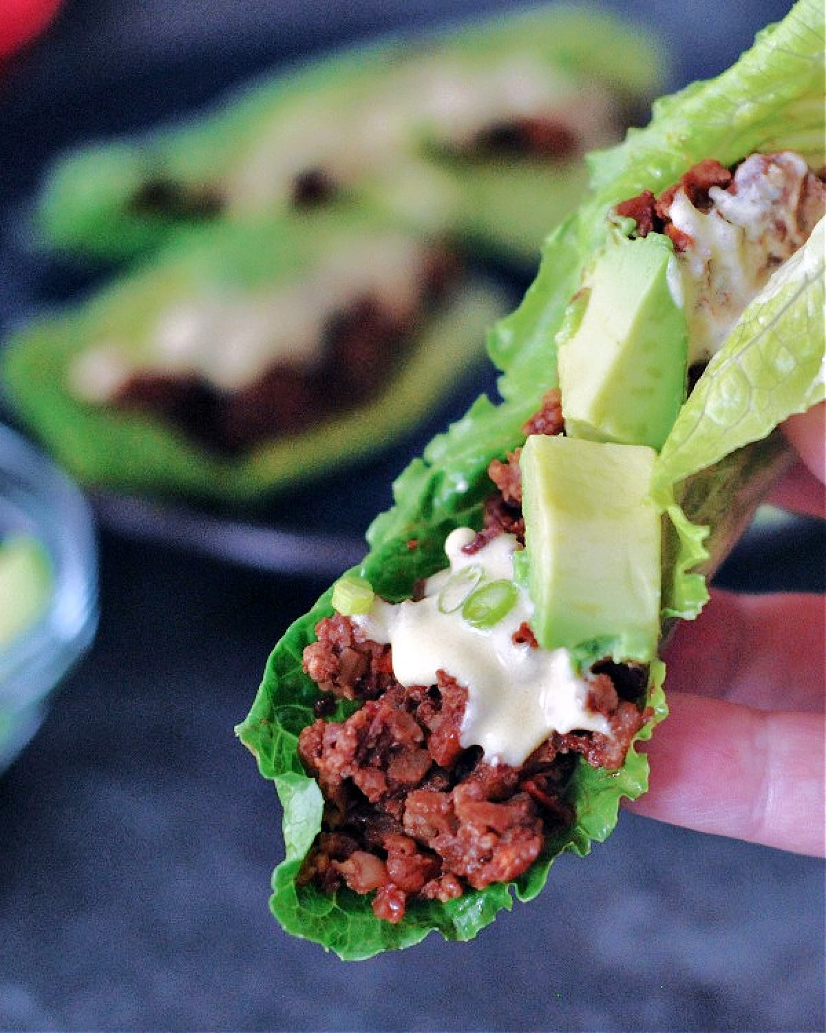 A hand holding a vegan chorizo sausage in lettuce wrap with avocado slices and dairy free cheese sauce.