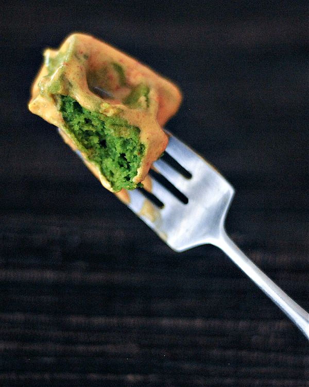 One bite of bright green spinach waffle on a fork, topped with orange colored sriracha whip cream.