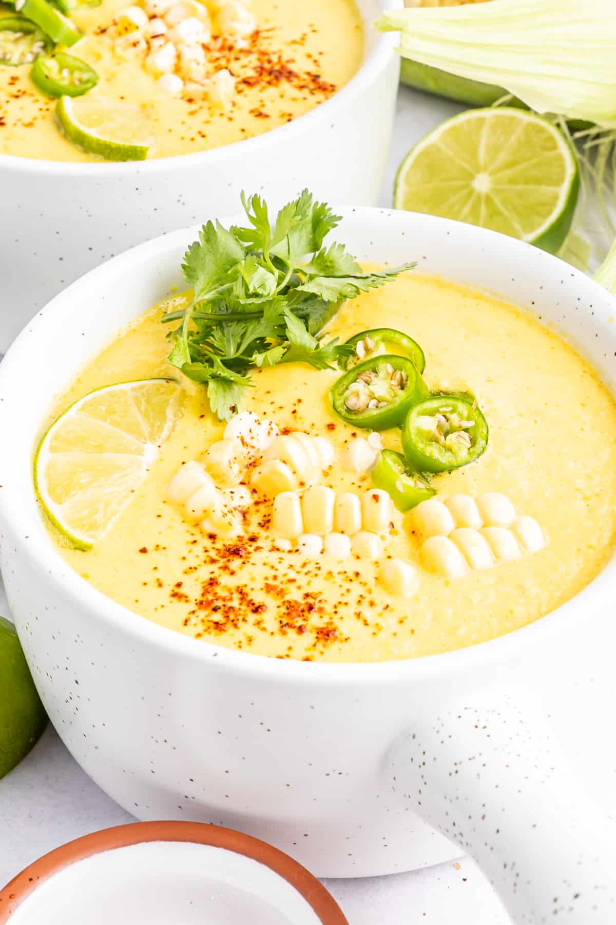 Bright yellow summer corn gazpacho in white bowls with handles. Garnished with corn pieces, sliced Serrano pepper, cilantro, and paprika. A full ear of corn sitting alongside bowls of soup, with extra line wedges, spoons, and a small bowl of paprika.