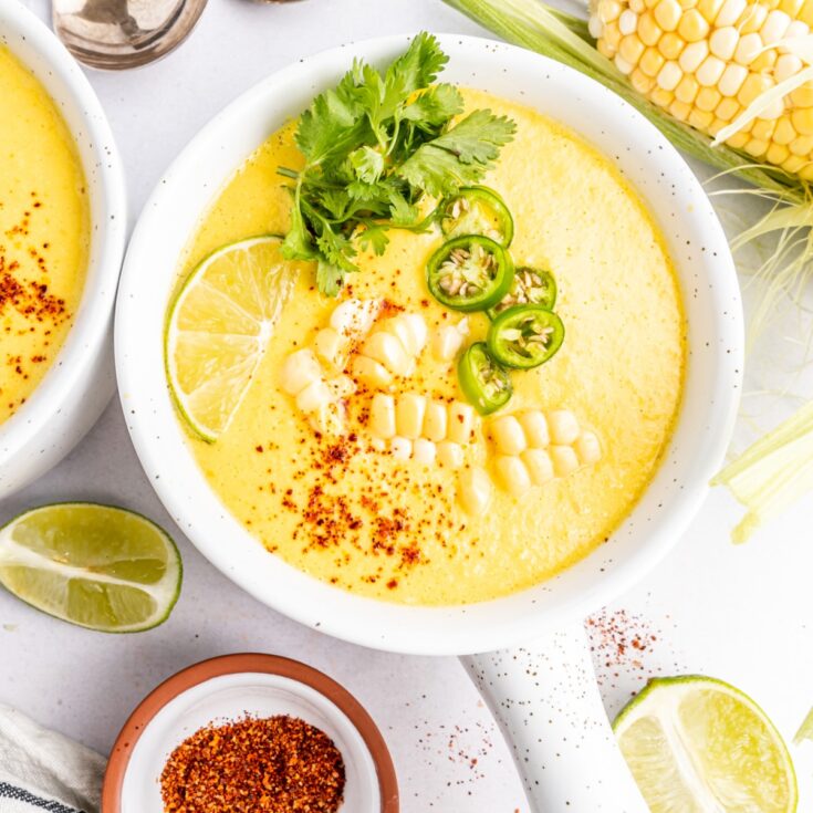 Overhead view of bright yellow summer corn gazpacho in white bowls with handles. Garnished with corn pieces, sliced Serrano pepper, cilantro, and paprika. A full ear of corn sitting alongside bowls of soup, with extra line wedges, spoons, and a small bowl of paprika.