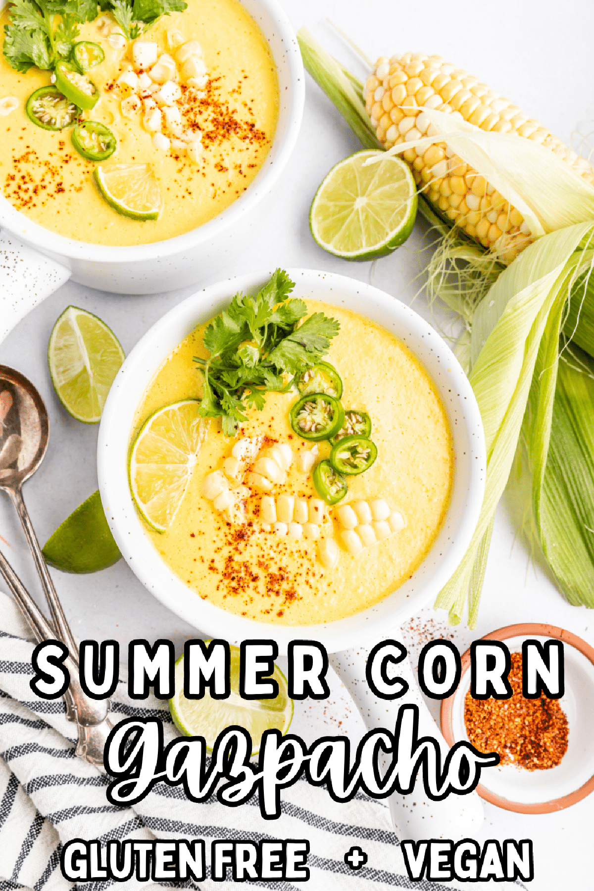 Overhead view of bright yellow summer corn gazpacho in white bowls with handles. Garnished with corn pieces, sliced Serrano pepper, cilantro, and paprika. A full ear of corn sitting alongside bowls of soup, with extra line wedges, spoons, and a small bowl of paprika.