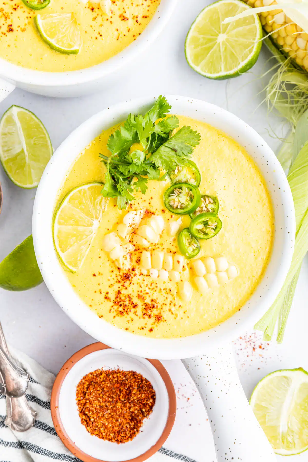 Overhead view of bright yellow summer corn gazpacho in white bowls with handles. Garnished with corn pieces, sliced Serrano pepper, cilantro, and paprika. A full ear of corn sitting alongside bowls of soup, with extra line wedges, spoons, and a small bowl of paprika.