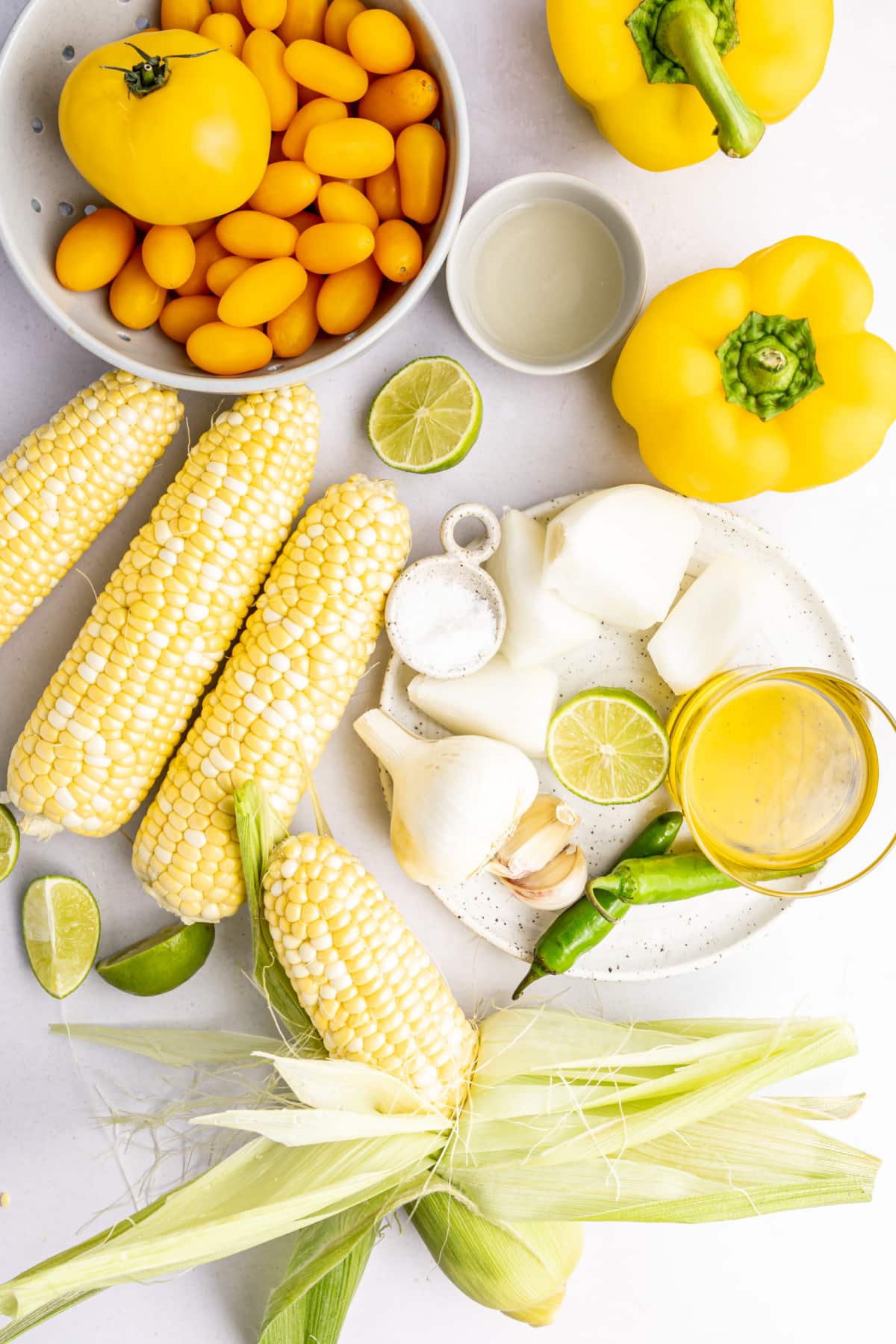 Overhead view of ingredients for summer corn gazpacho: yellow tomatoes, yellow bell pepper, fresh corn on the cob, limes, garlic bulbs, Serrano peppers