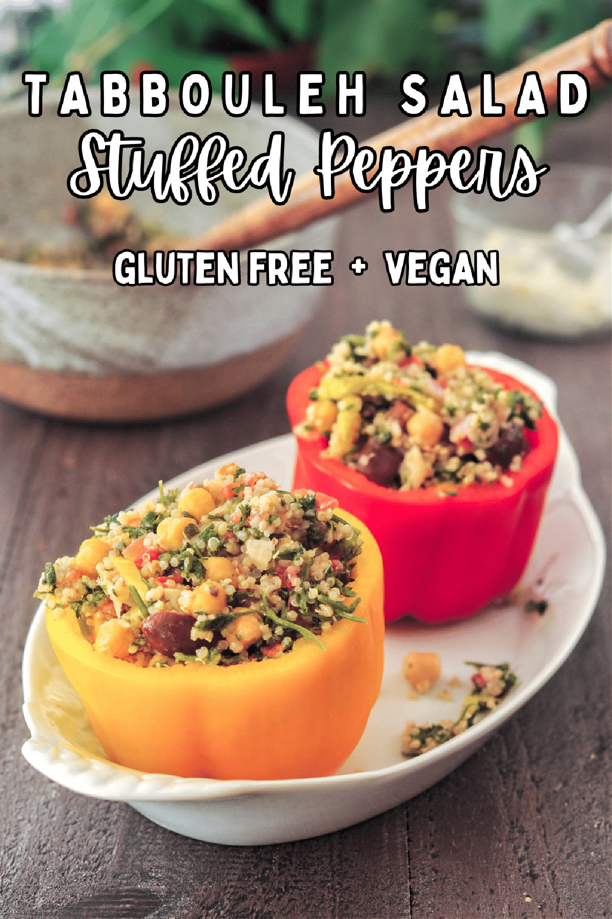 An orange and a red bell pepper with the tops cut off, filled with a chickpea tabbouleh salad. Peppers are sitting in a white oval dish on a dark wood table, with a grey bowl filled with more tabbouleh salad.