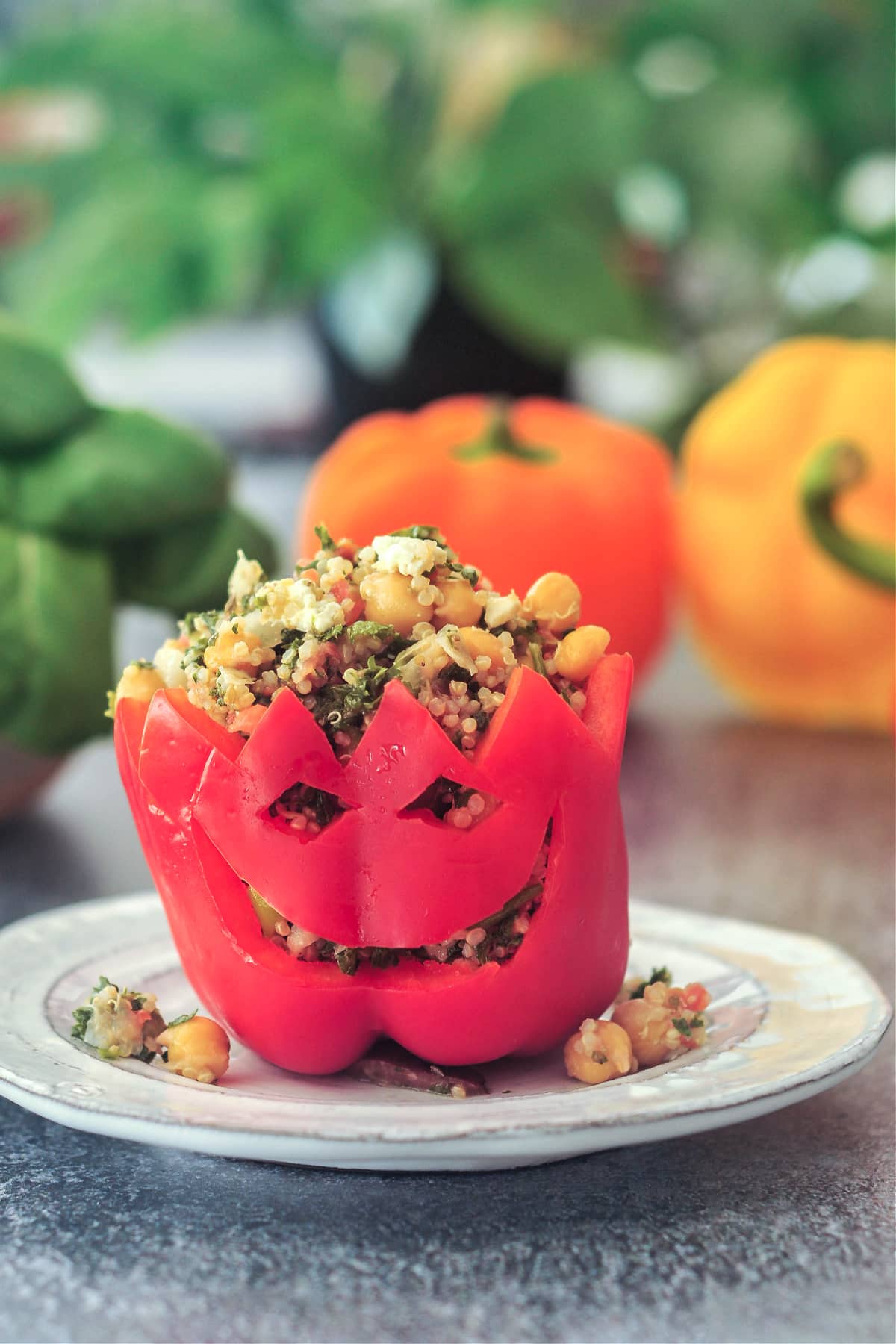 A red bell pepper filled with chickpea tabbouleh salad sits upright on a rustic white plate. The bell pepper has a jack o lantern face carved into the side. An orange and a yellow bell pepper and a bowl of greens sits in background, blurred slightly.