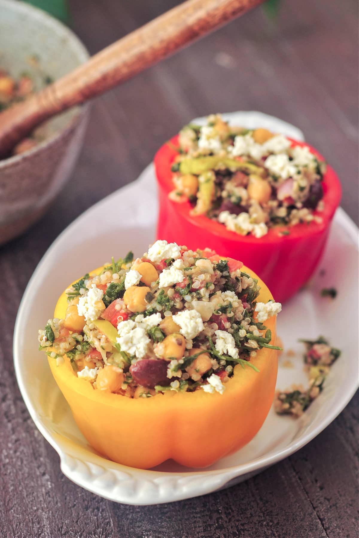 An orange and a red bell pepper with the tops cut off, filled with a chickpea tabbouleh salad. Peppers are sitting in a white oval dish on a dark wood table, with a grey bowl filled with more tabbouleh salad.
