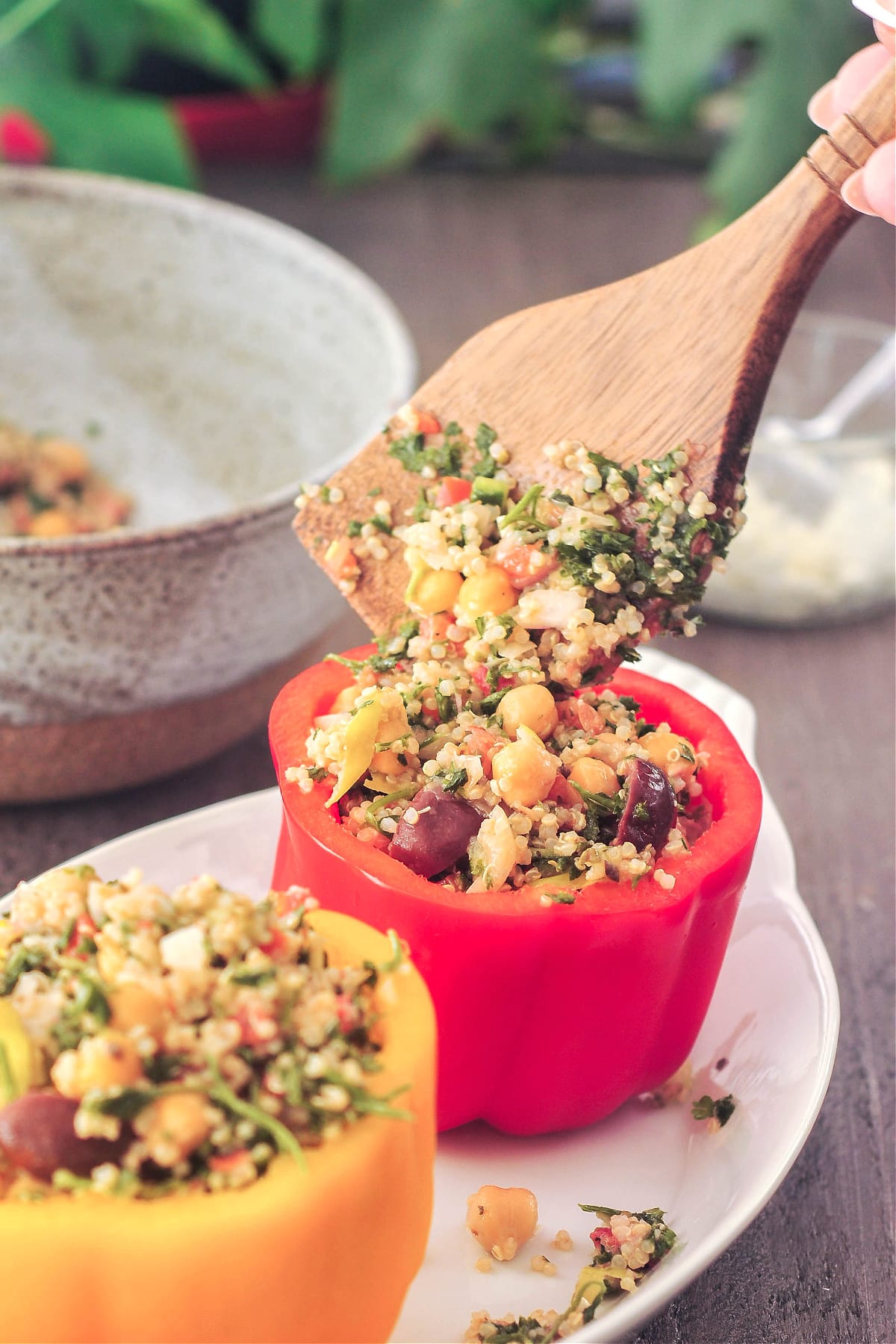 An orange and a red bell pepper with the tops cut off. A flat wooden spoon is filling the peppers with a chickpea tabbouleh salad. Peppers are sitting in a white oval dish on a dark wood table, with a grey bowl filled with more tabbouleh salad.