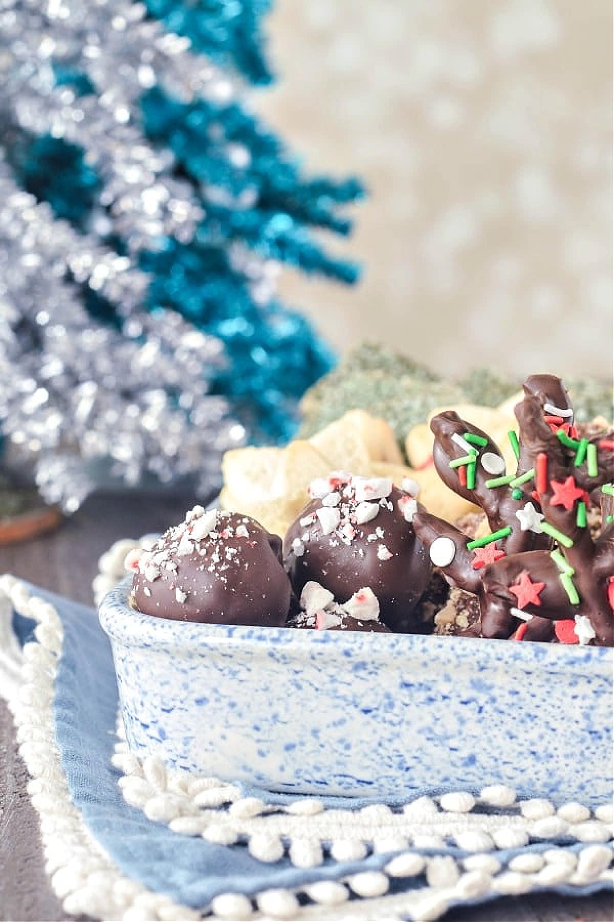 A blue and white dish of holiday treats, including chocolate coated truffles with candy cane pieces, pretzel reindeer, shortbread cookies. A silver and teal foil Christmas tree sits in background.