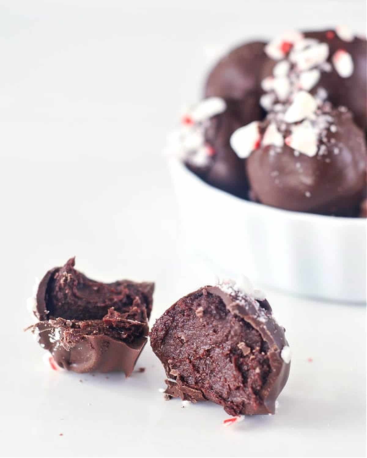 A small ramekin of chocolate coated truffles with candy cane pieces on top sits on a white tabletop. One single truffle sits outside the bowl, cut in half to show chocolate inside.