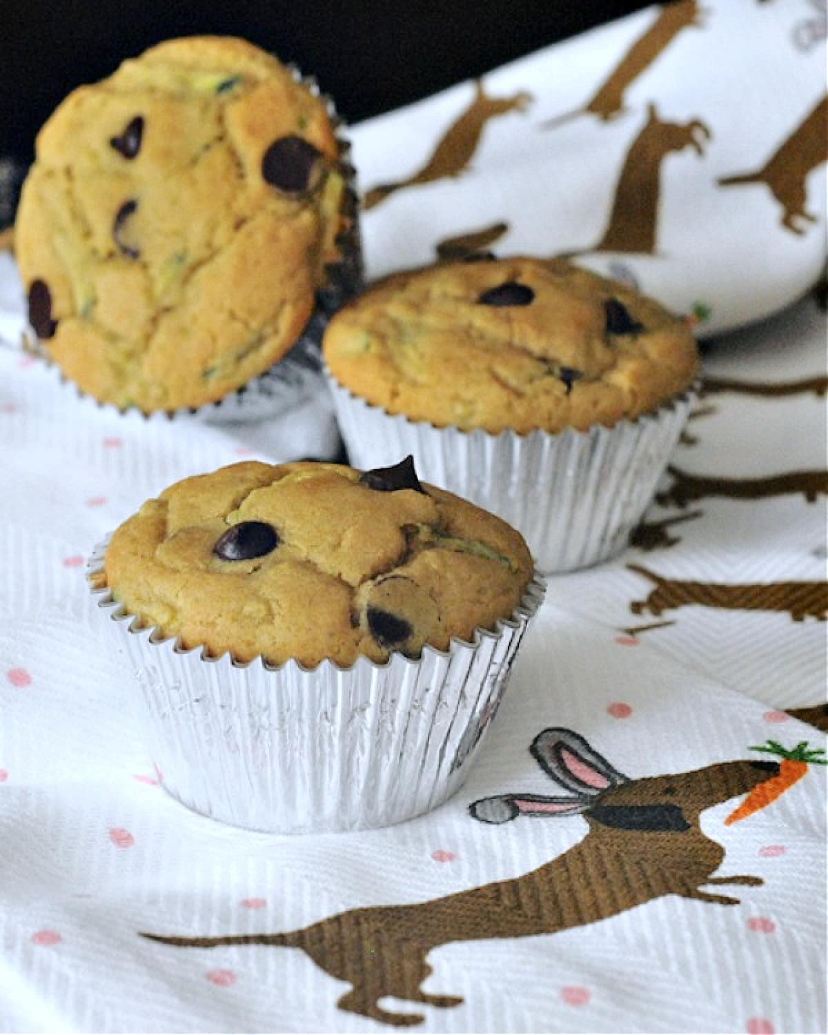 Three chocolate chip zucchini muffins sitting on a white cloth napkin printed with brown dachshund dogs on it.