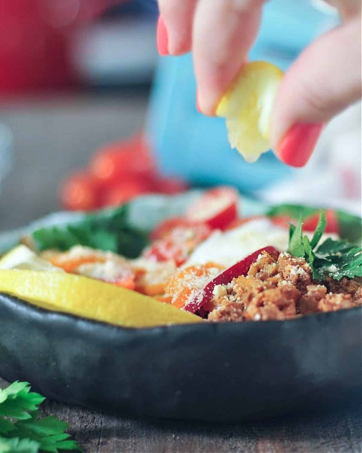 Close up of a hand squeezing lemon over a deconstructed stuffed pepper bowl (spinach and greens, vegan ground beef, cherry tomatoes, bell pepper slices, green onion and cilantro).