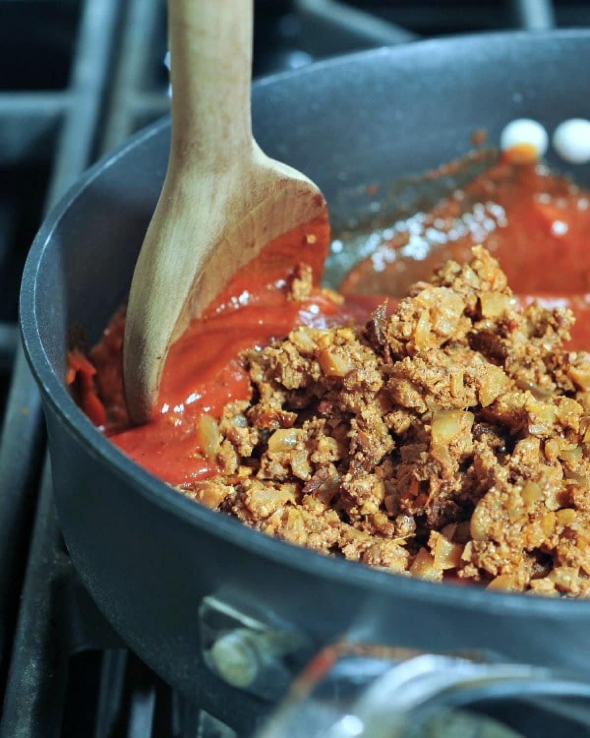 Vegan ground beef being stirred into red sauce in a skillet on a stove top.