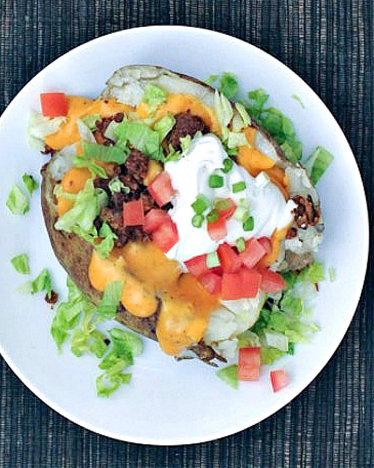 Overhead view of vegan ground beef in a taco baked potato, with shredded lettuce, diced tomato, sour cream, green onion, and cheese sauce.