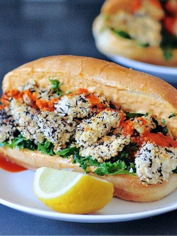 Vegan oyster mushroom po boy sandwich on a white plate, dressed with kale, spicy garlic sauce, and lemon wedges.