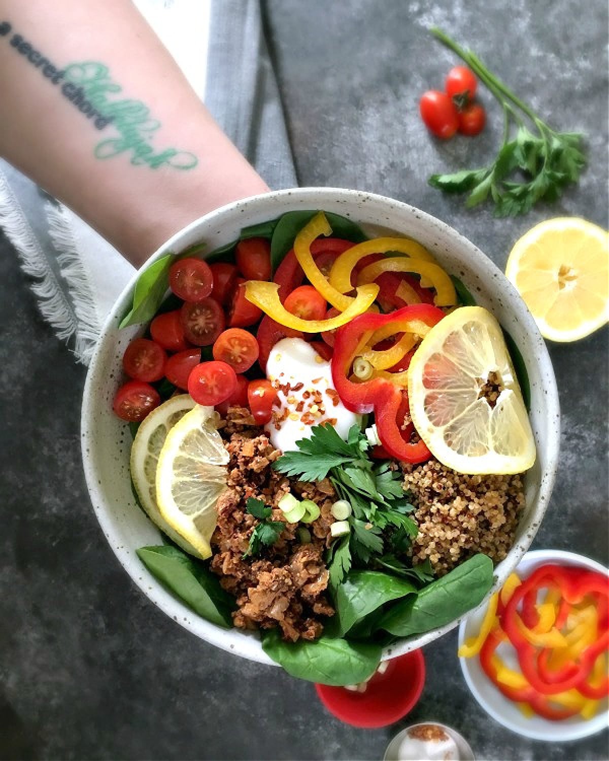 Overhead view of a tattooed arm and hand holding a deconstructed stuffed pepper bowl (spinach and greens, vegan ground beef, cherry tomatoes, bell pepper slices, green onion and cilantro).