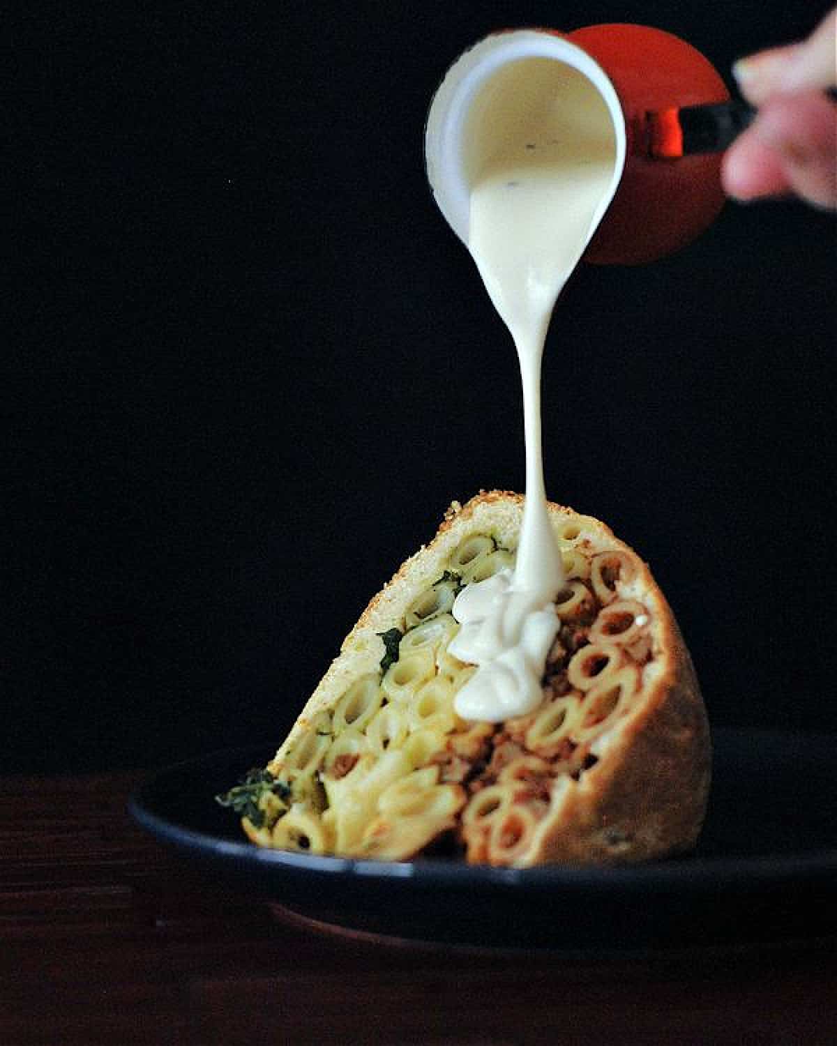 A hand pouring Alfredo sauce over a single serving of timpano against a black background. Timpano is a large pastry dome filled with pasta: a red marinara layer of penne pasta on top, a middle layer of white Alfredo pasta, and a bottom layer of green pesto pasta.