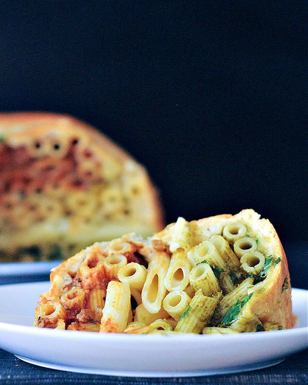 A single serving of timpano on a plate in front of the platter of a full timpano. Timpano is a large pastry dome filled with pasta: a red marinara layer of penne pasta on top, a middle layer of white Alfredo pasta, and a bottom layer of green pesto pasta.