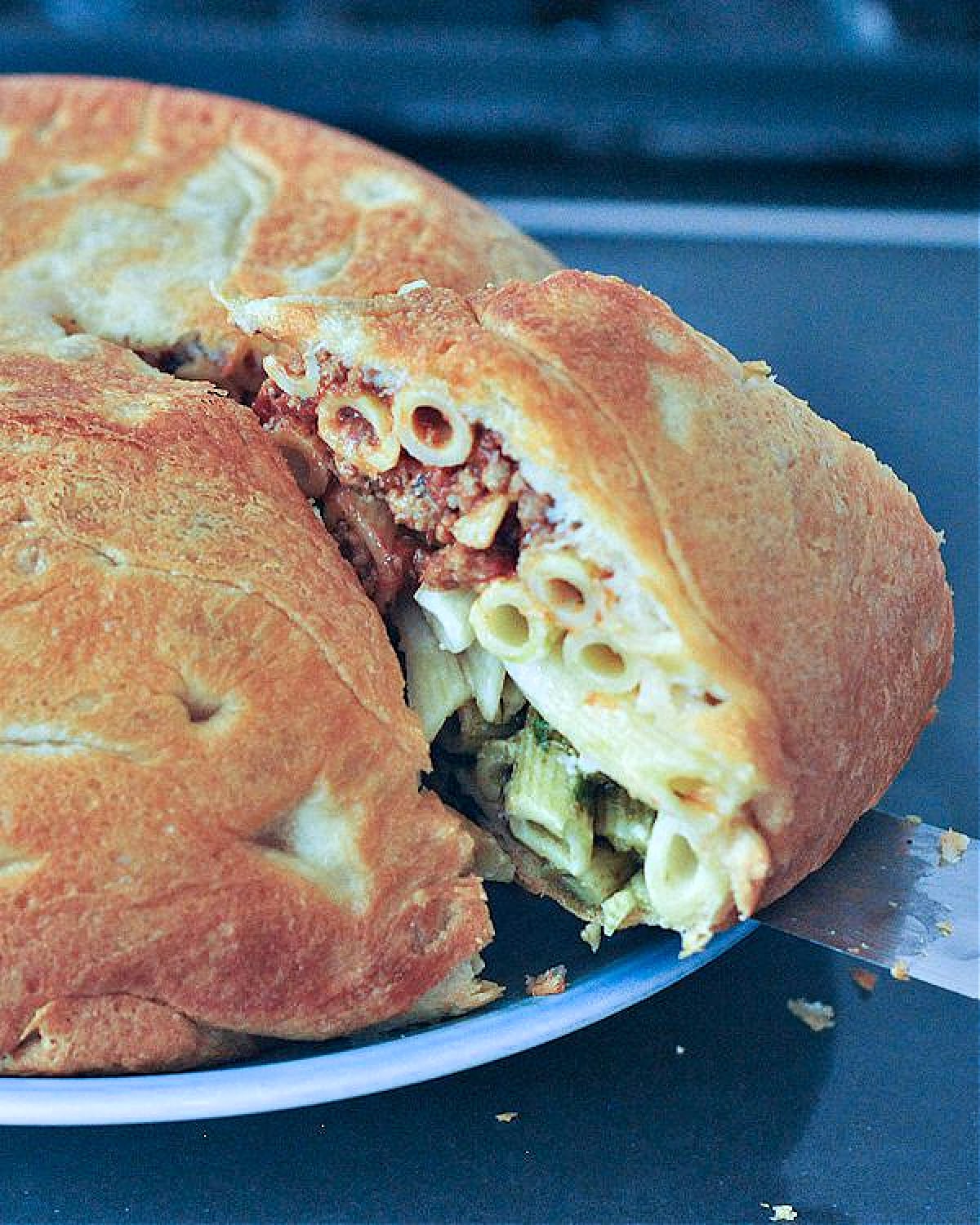 A single serving of timpano being cut and removed from a full timpano. Timpano is a large pastry dome filled with pasta: a red marinara layer of penne pasta on top, a middle layer of white Alfredo pasta, and a bottom layer of green pesto pasta.