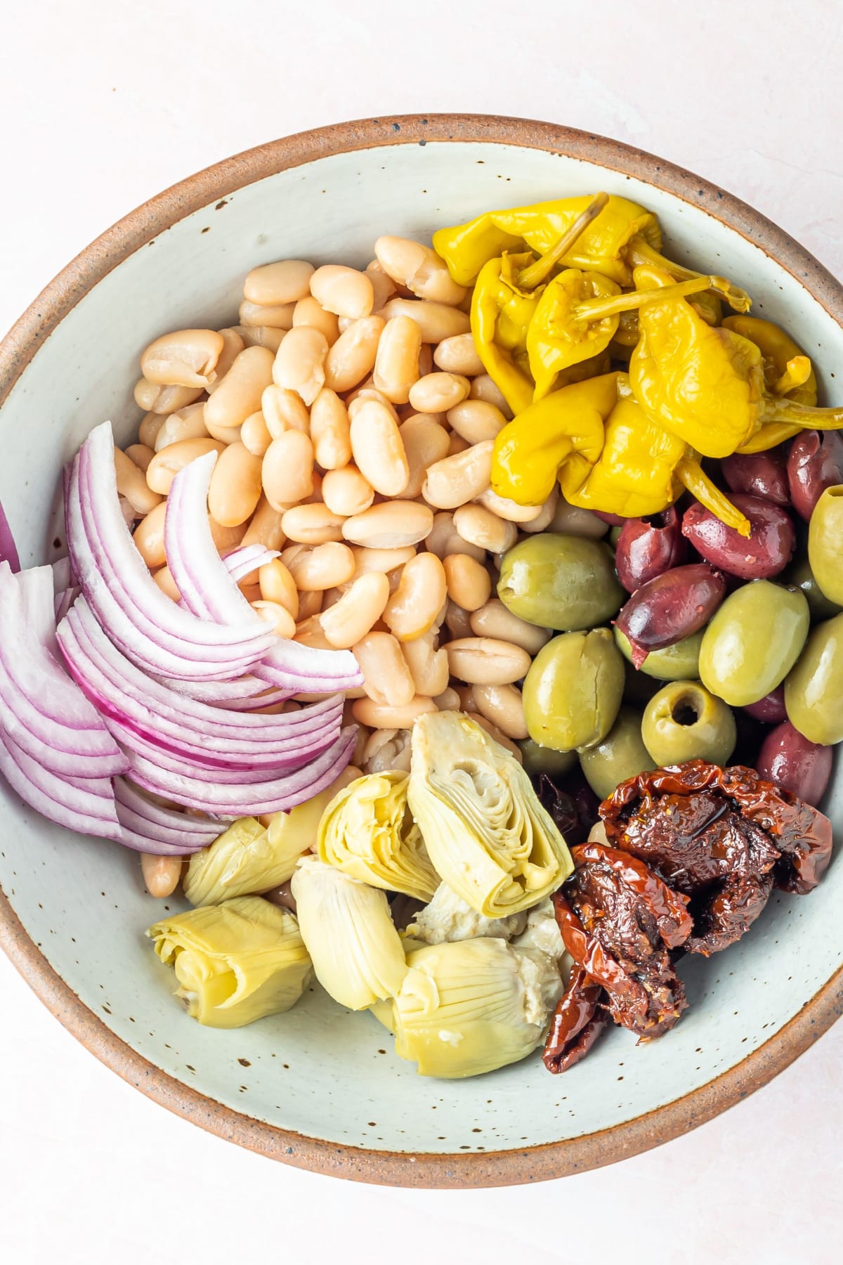 Overhead view of a bowl of white bean antipasto salad before mixing, which includes black kalamata and green castelvetrano olives, white beans, sun dried tomatoes, sliced artichoke hearts, sliced red onion, pepperoncini peppers, herbs and spices.