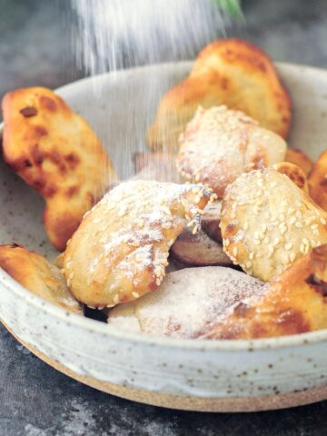 A shallow grey bowl of air fried banana fritters being dusted with powdered sugar.