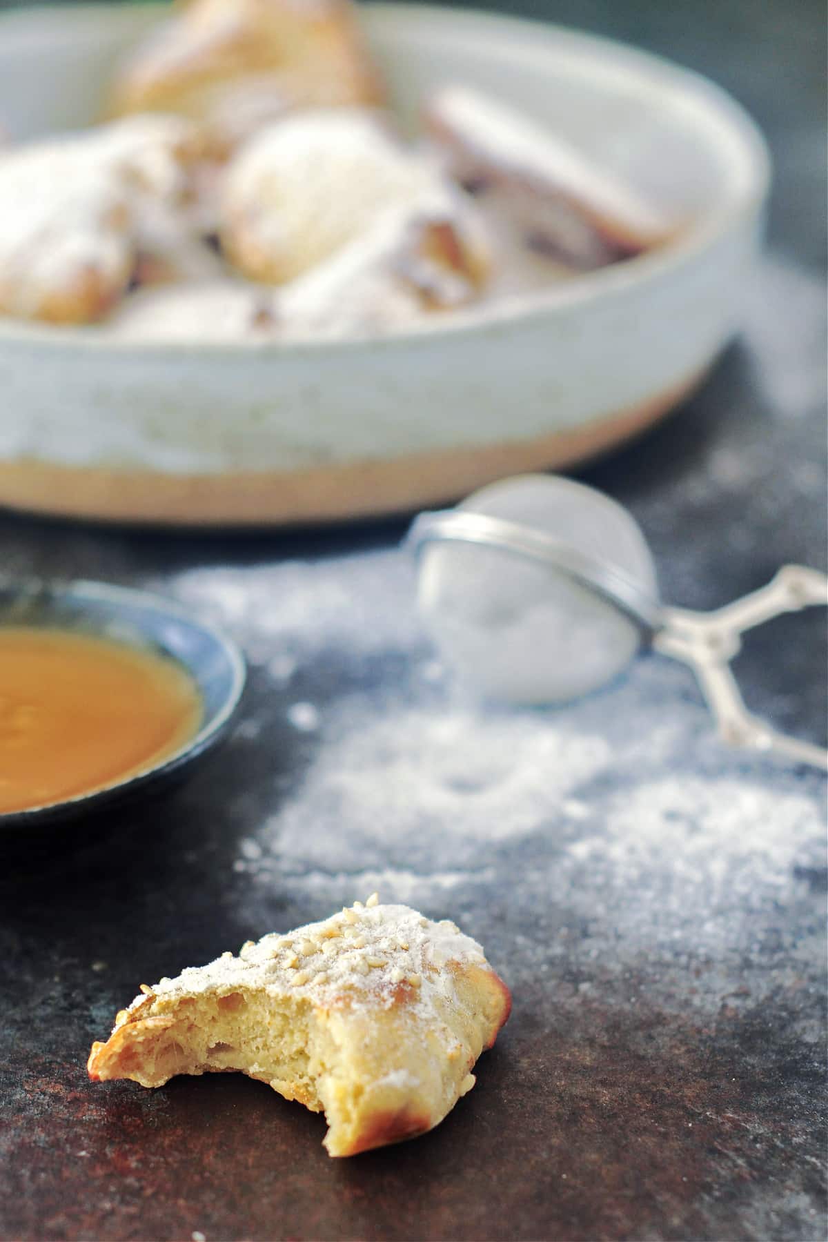 A banana fritter with a bite out of it sits next to a bowl of fritters and a bowl of caramel sauce. Powdered sugar dusts the surface around the bowl, and a mesh strainer sits in the powdered sugar dust.